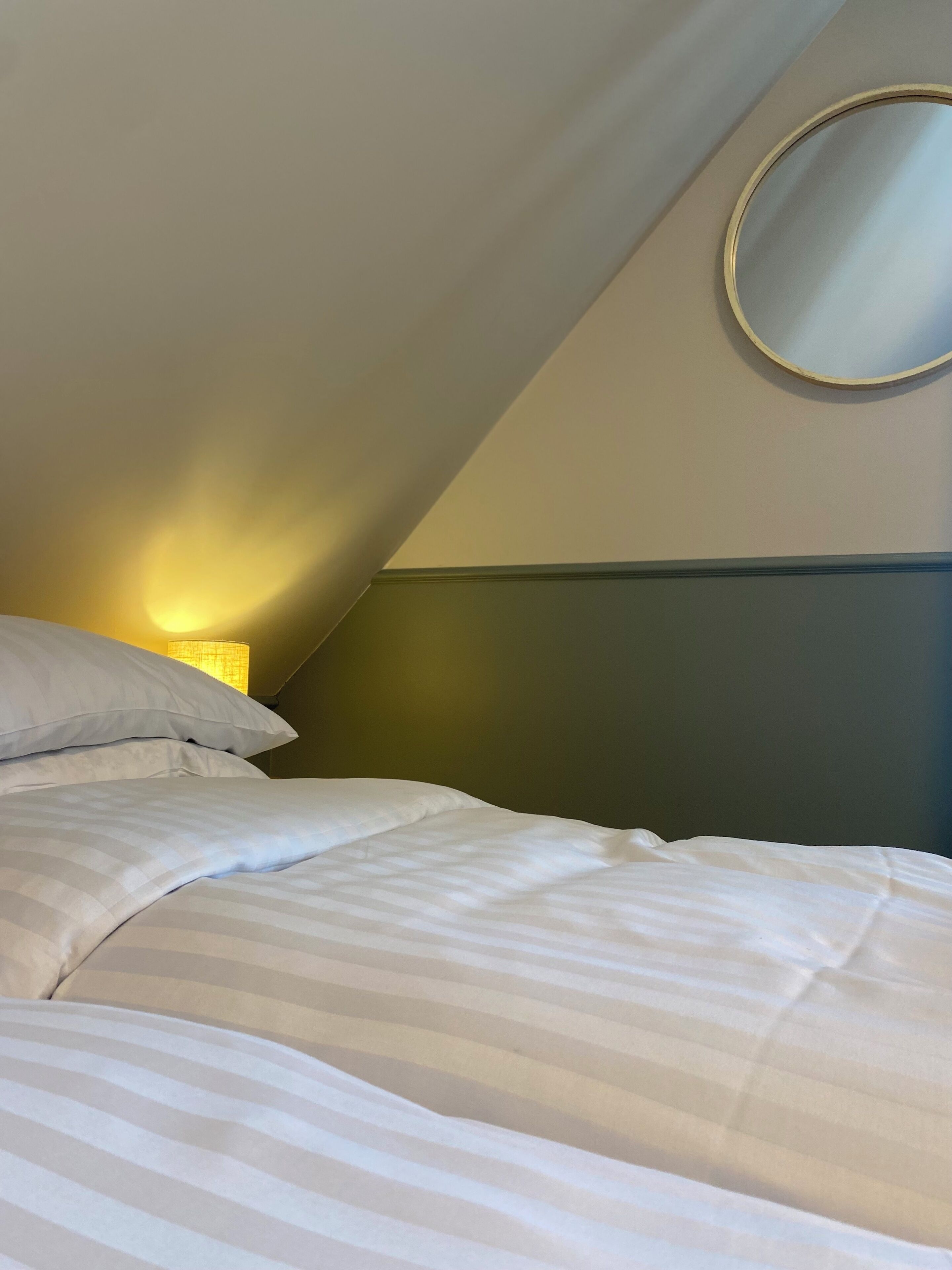 A bedroom with a bed, pillows, a nightstand, a lamp, and a round mirror on the wall.