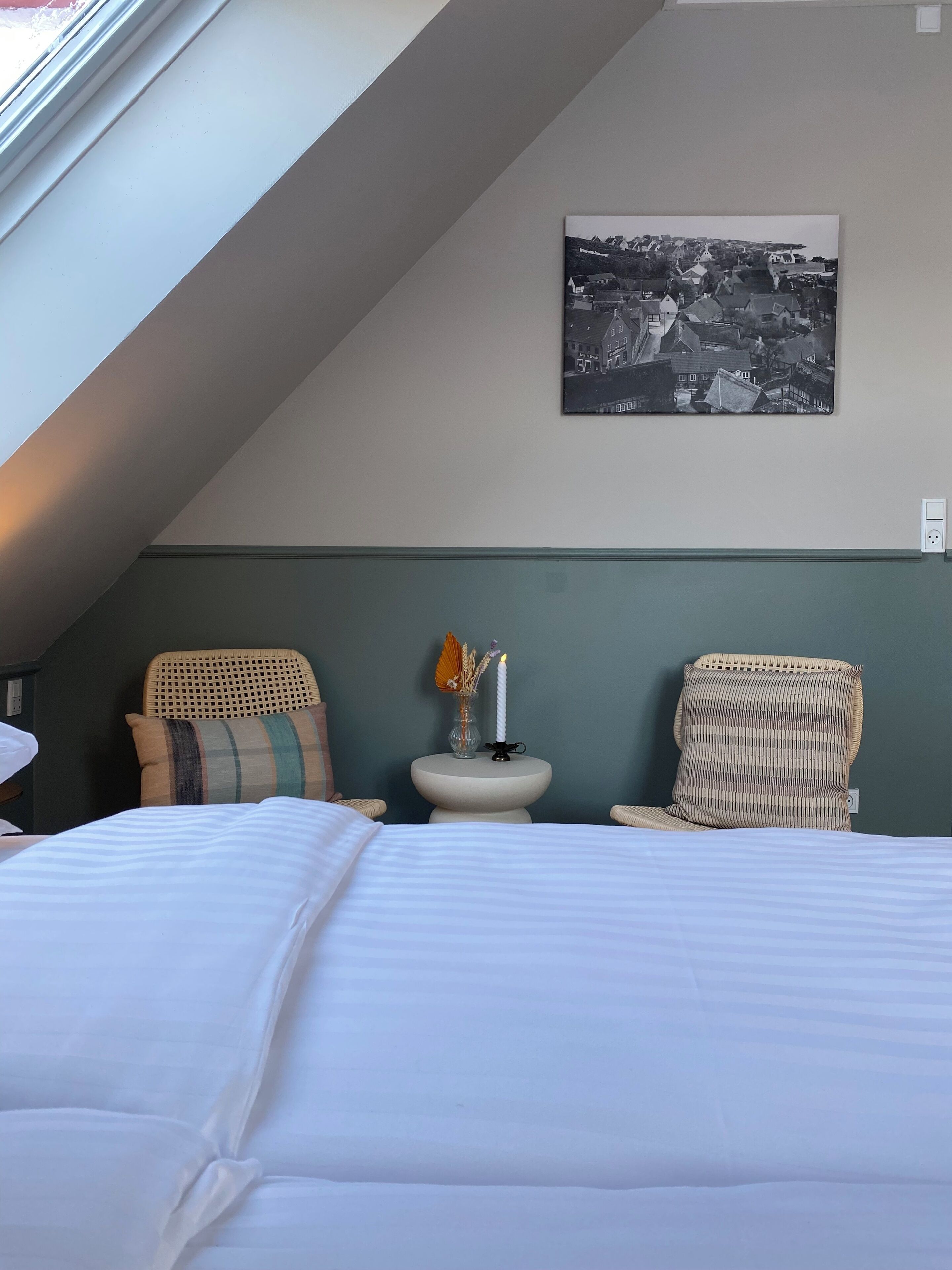 Une chambre à coucher avec un lit, deux fauteuils en osier, une petite table avec une bougie et une photographie en noir et blanc accrochée au mur.