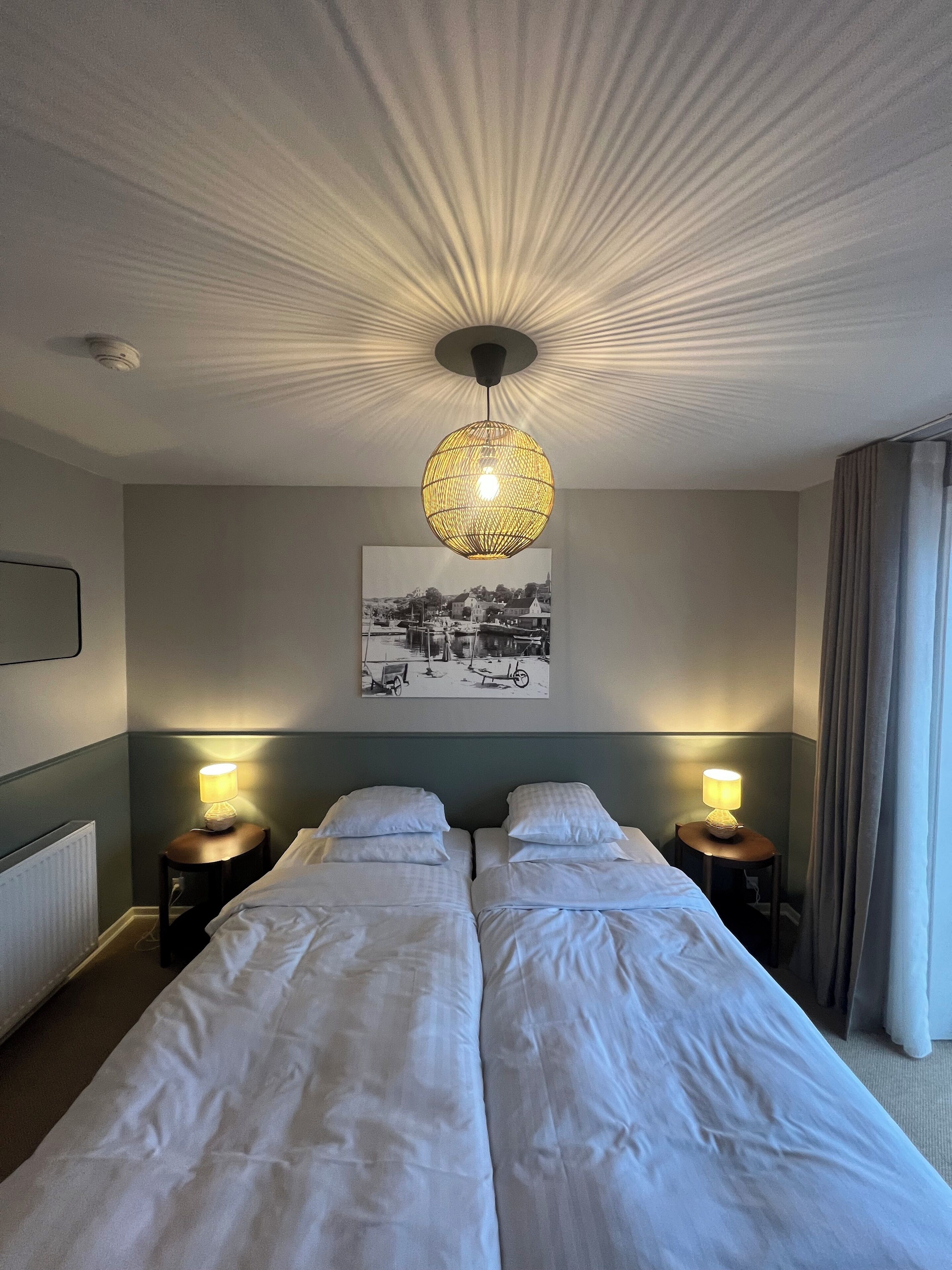A hotel room with two beds, a ceiling light fixture, a wall-mounted picture, and a window with curtains.