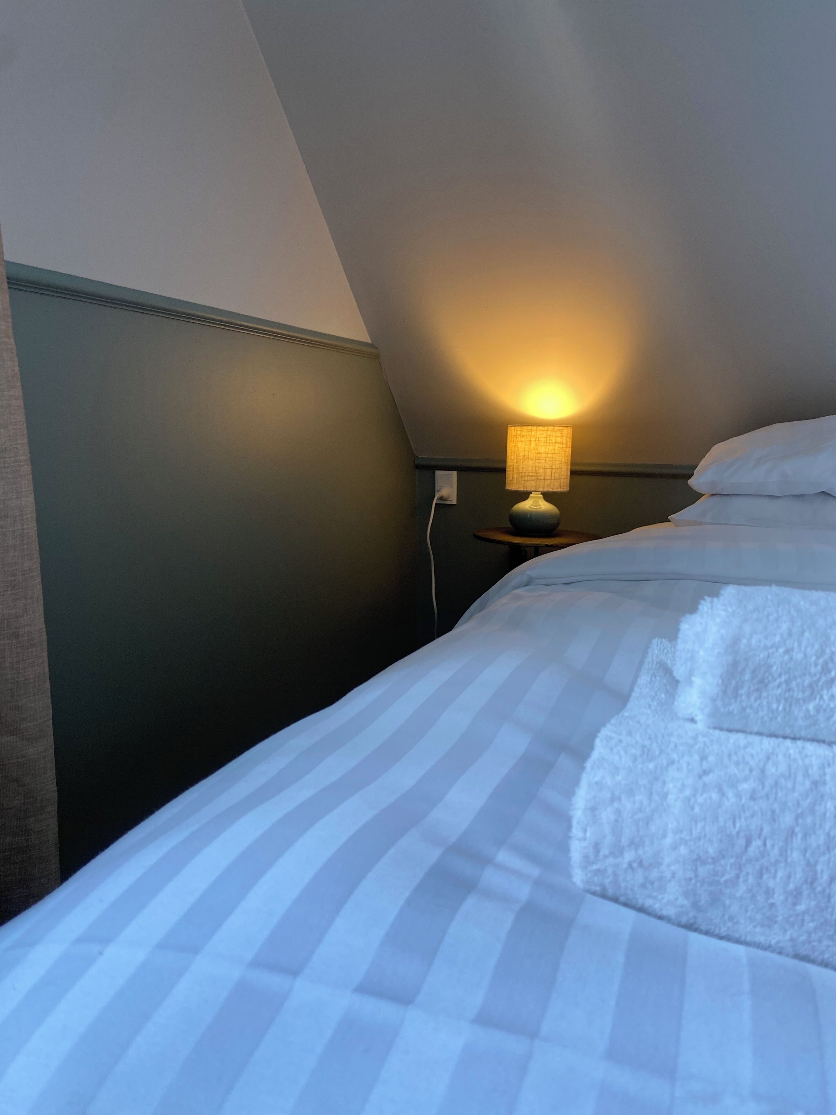 A neatly made bed with striped bedding and a white pillow, a bedside lamp, and a power outlet on a dark wall in a hotel room.