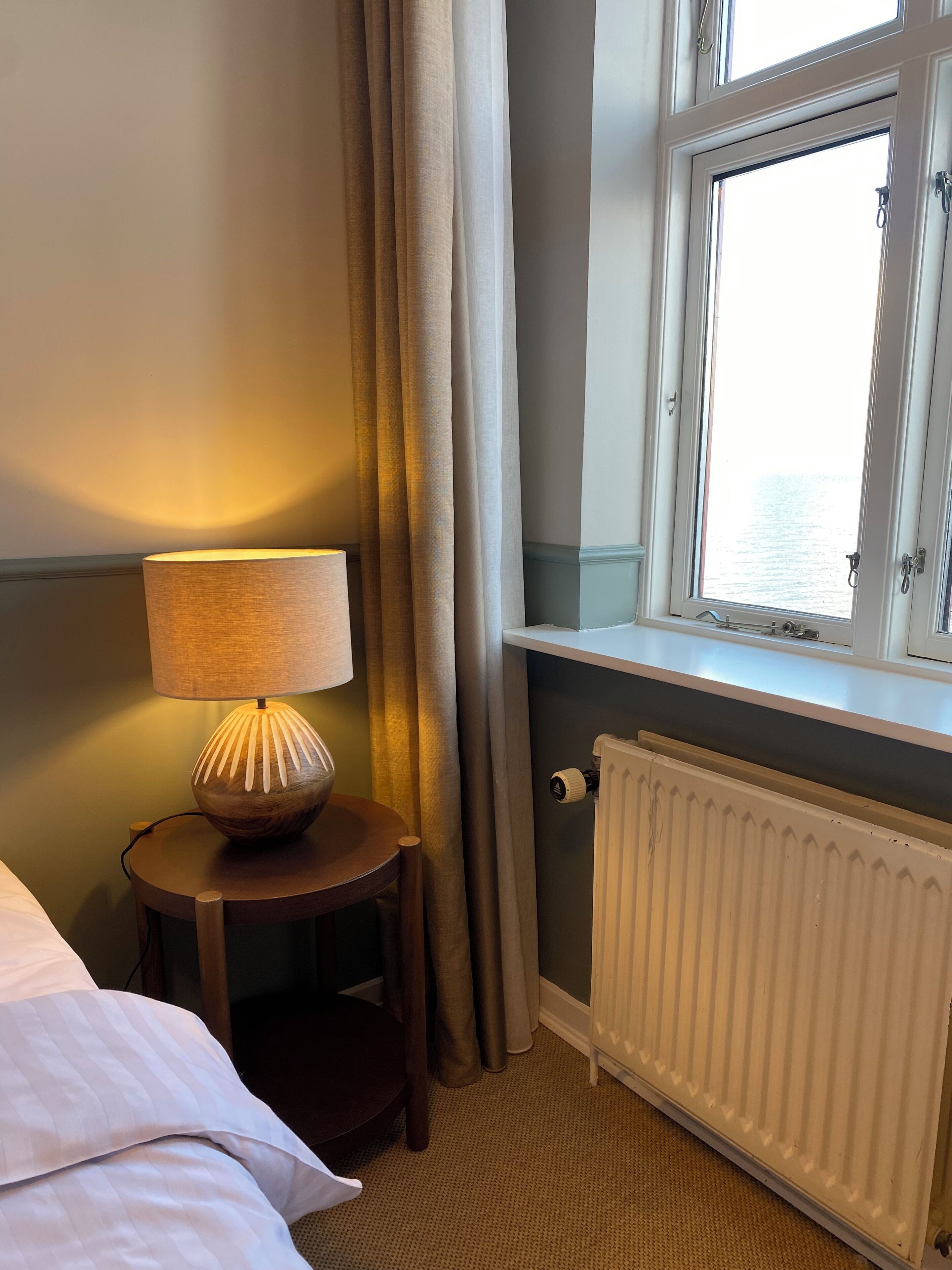Et hotelværelse med seng, sengetæppe, lampe, vindue med udsigt og radiator.