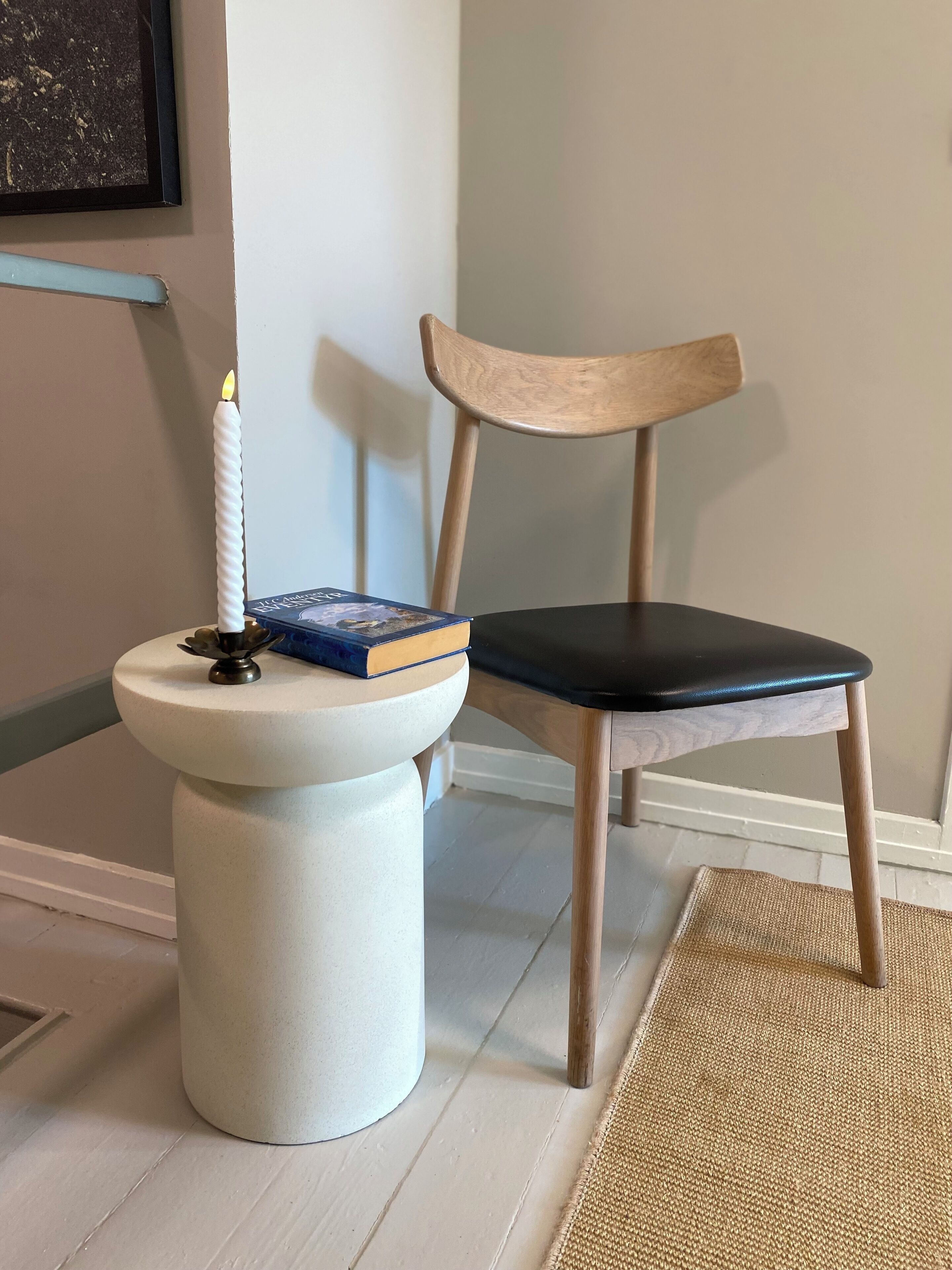 Une petite table blanche sur pied, ornée d’une bougie allumée, d’un livre et d’une chaise noire avec un dossier en bois.