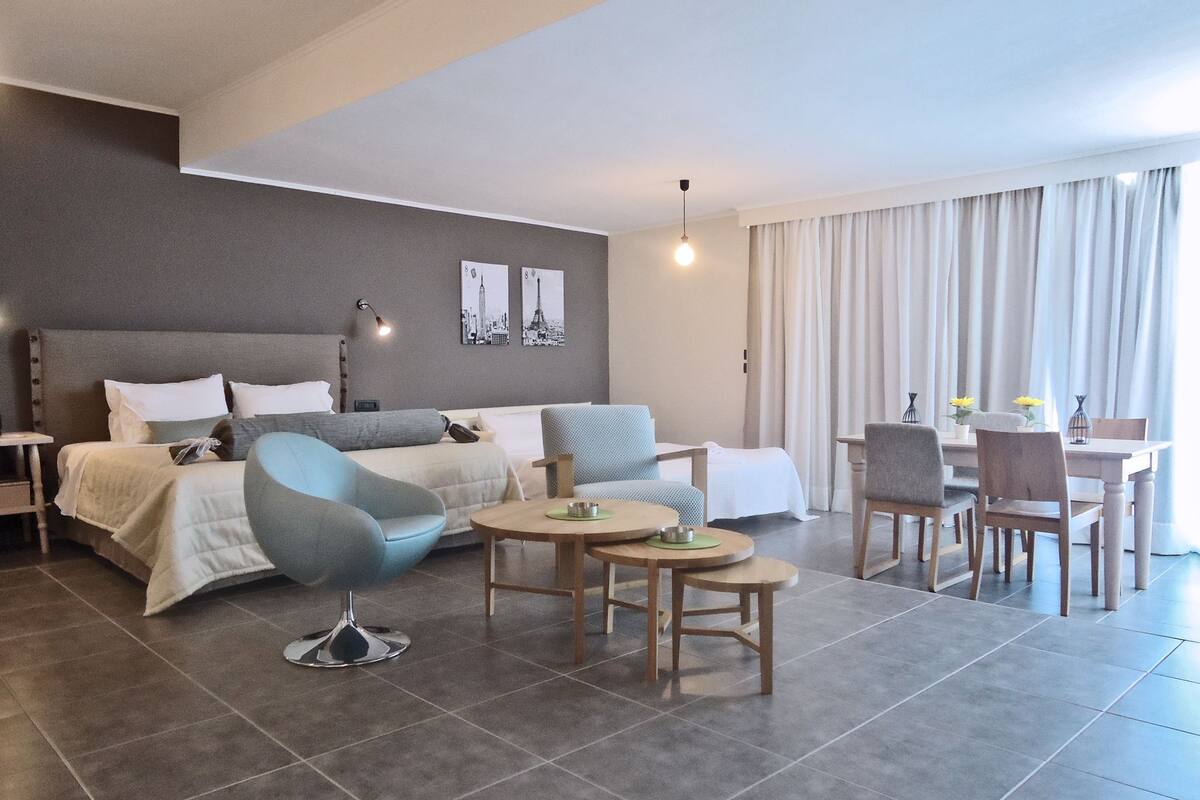 A modern hotel room with a bed, a sofa, a dining table, and chairs.