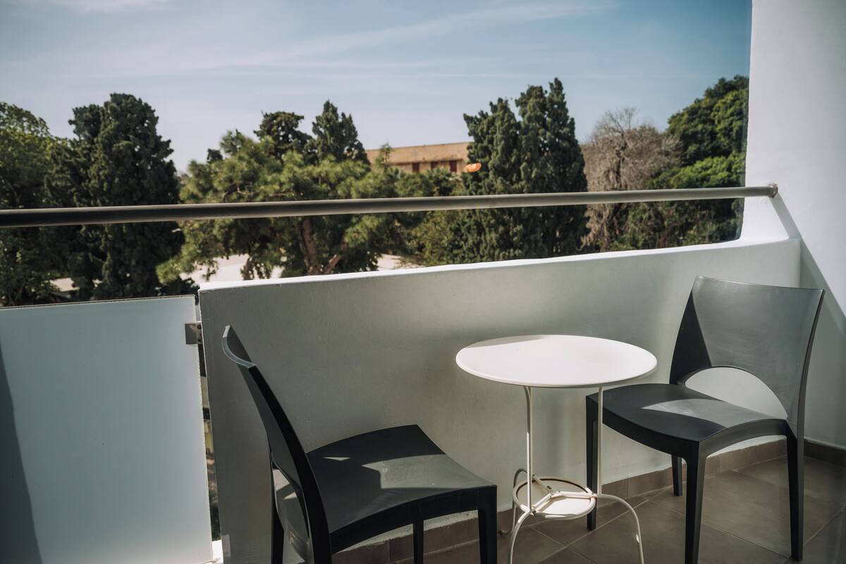 A balcony with two chairs and a small table, overlooking a view of trees and a building.