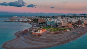 An aerial view of a coastal city with a beach, buildings, and a cruise ship docked nearby. - Amphitryon City Hotel (Rhodes)