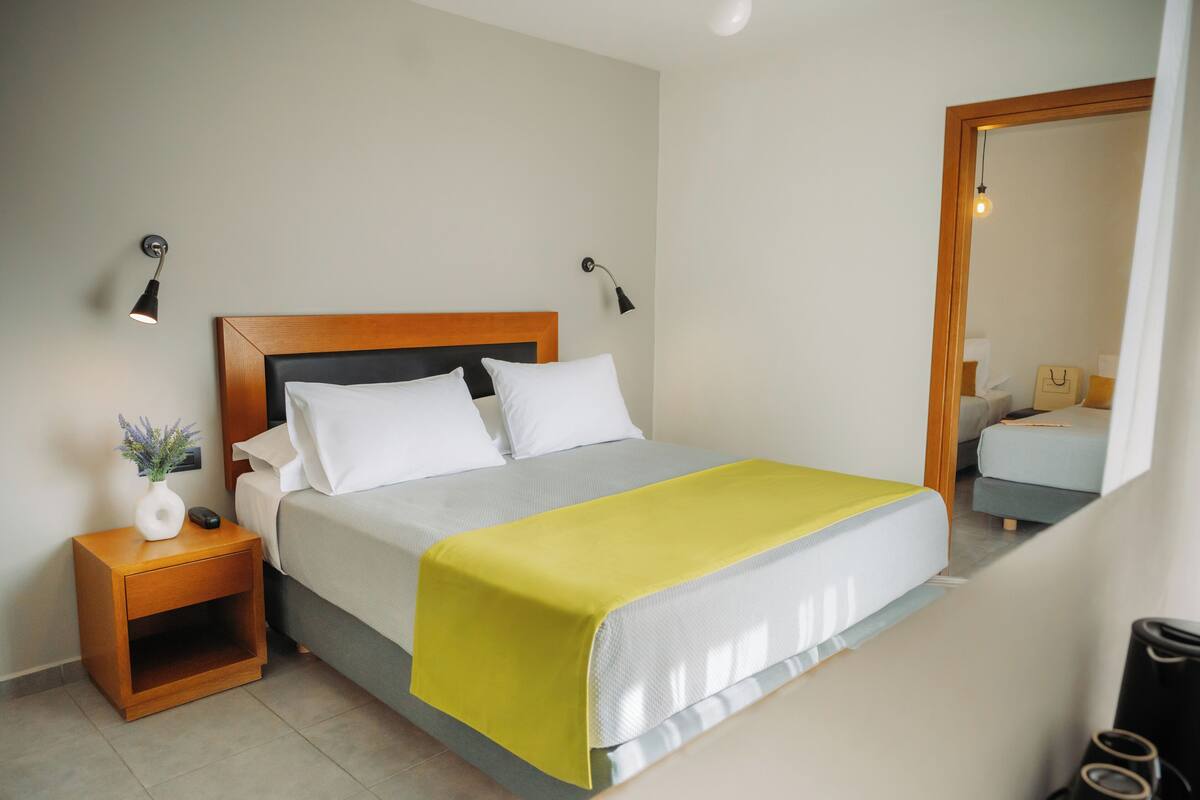 A neatly made bed with a wooden headboard, a bedside table with a vase and a remote control, and two wall-mounted lamps.