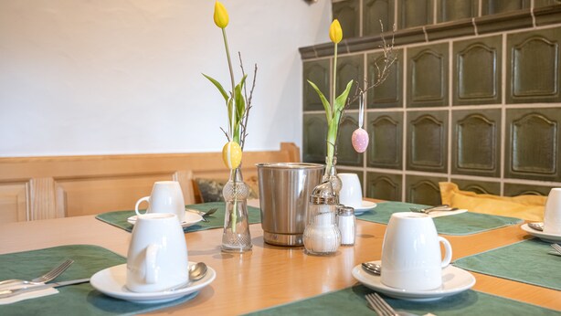 A dining table set with white cups, saucers, and a variety of tableware. There are two vases with flowers and a salt and pepper set on the table. - Hotel Neuwirt (Sauerlach)