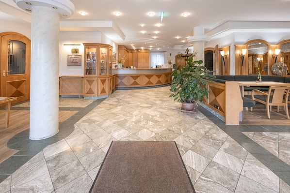 A spacious hotel lobby with a marble floor, wooden reception desk, and a large potted plant. - Hotel Neuwirt (Sauerlach)
