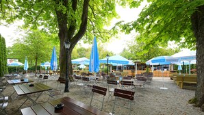 An outdoor seating area with tables and benches, surrounded by trees and covered by blue umbrellas. - Hotel Neuwirt (Sauerlach)