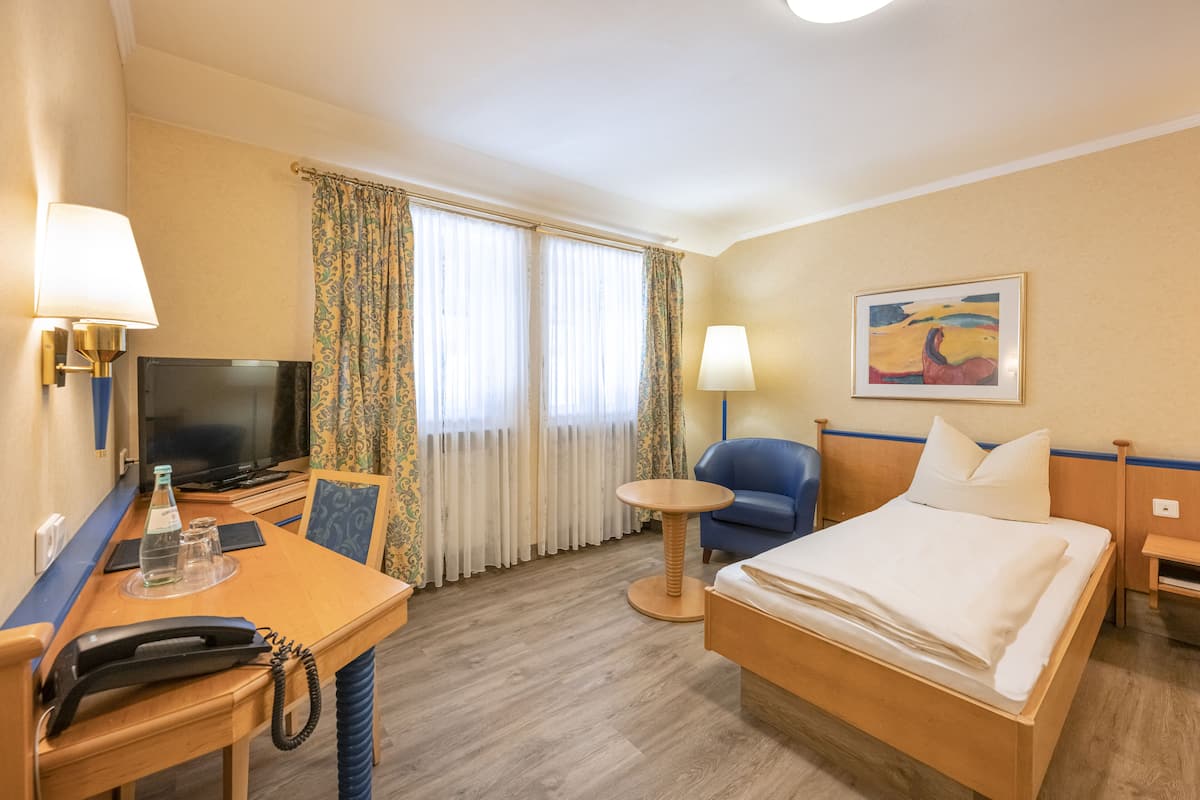 Habitación de hotel con cama, escritorio con lámpara, silla, televisión y un cuadro en la pared.