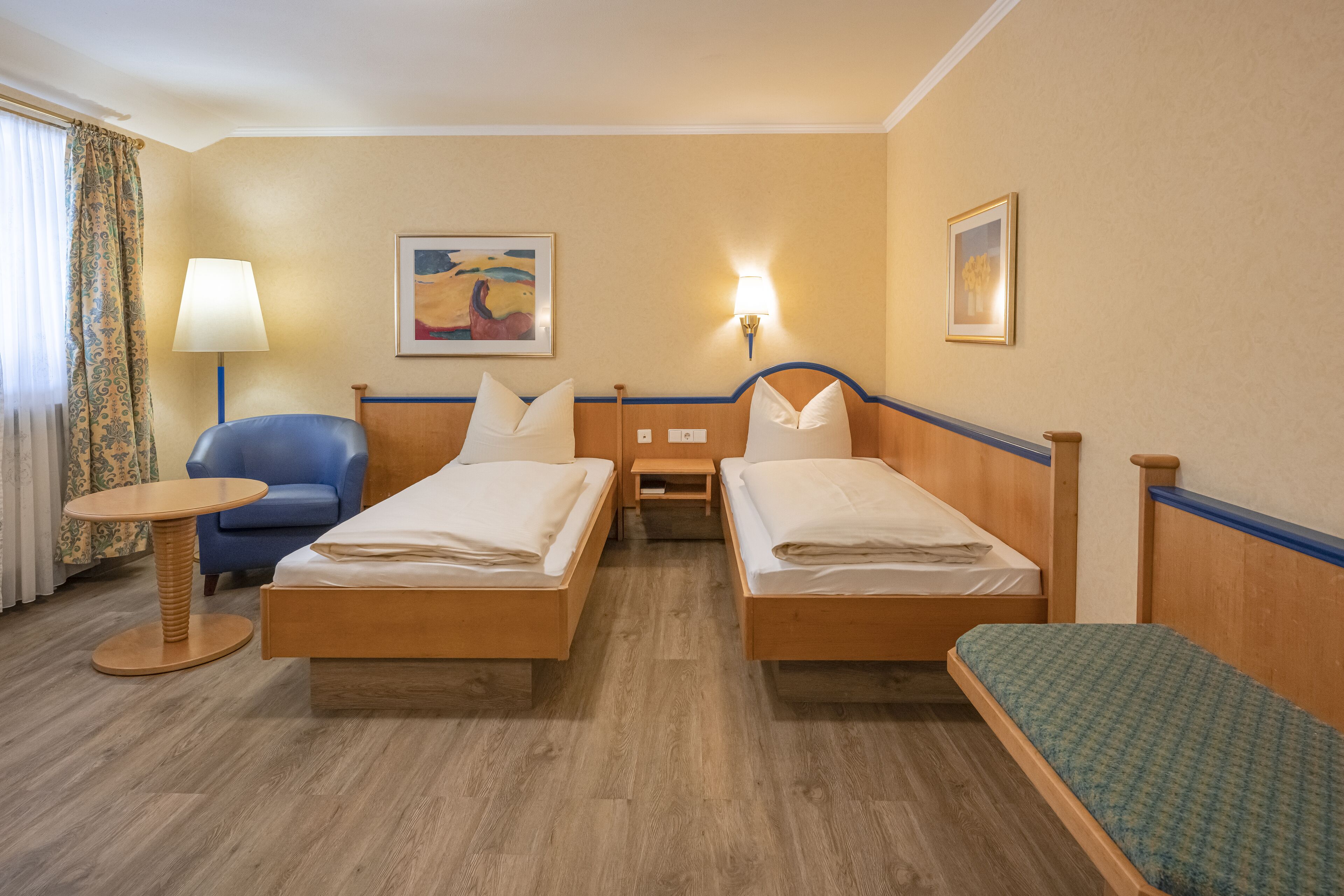 Une chambre d’hôtel avec deux lits en bois, une chaise bleue, une petite table ronde et une lampe.
