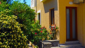 A building with a yellow facade, a stone staircase, and a small garden with yellow flowers and red roses. - Katerini Hotel Apartments (Rethymno)