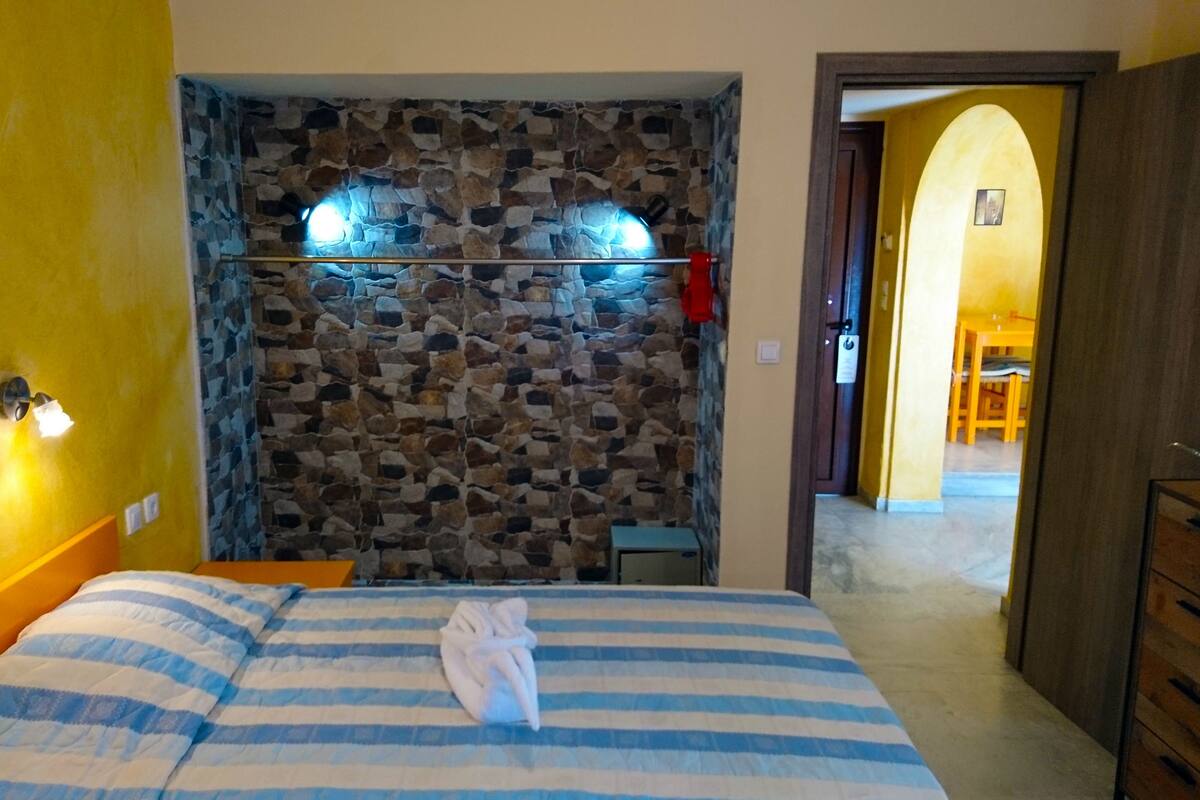 A bedroom with a stone wall, a bed with striped bedding, a nightstand, and a wooden door leading to another room.