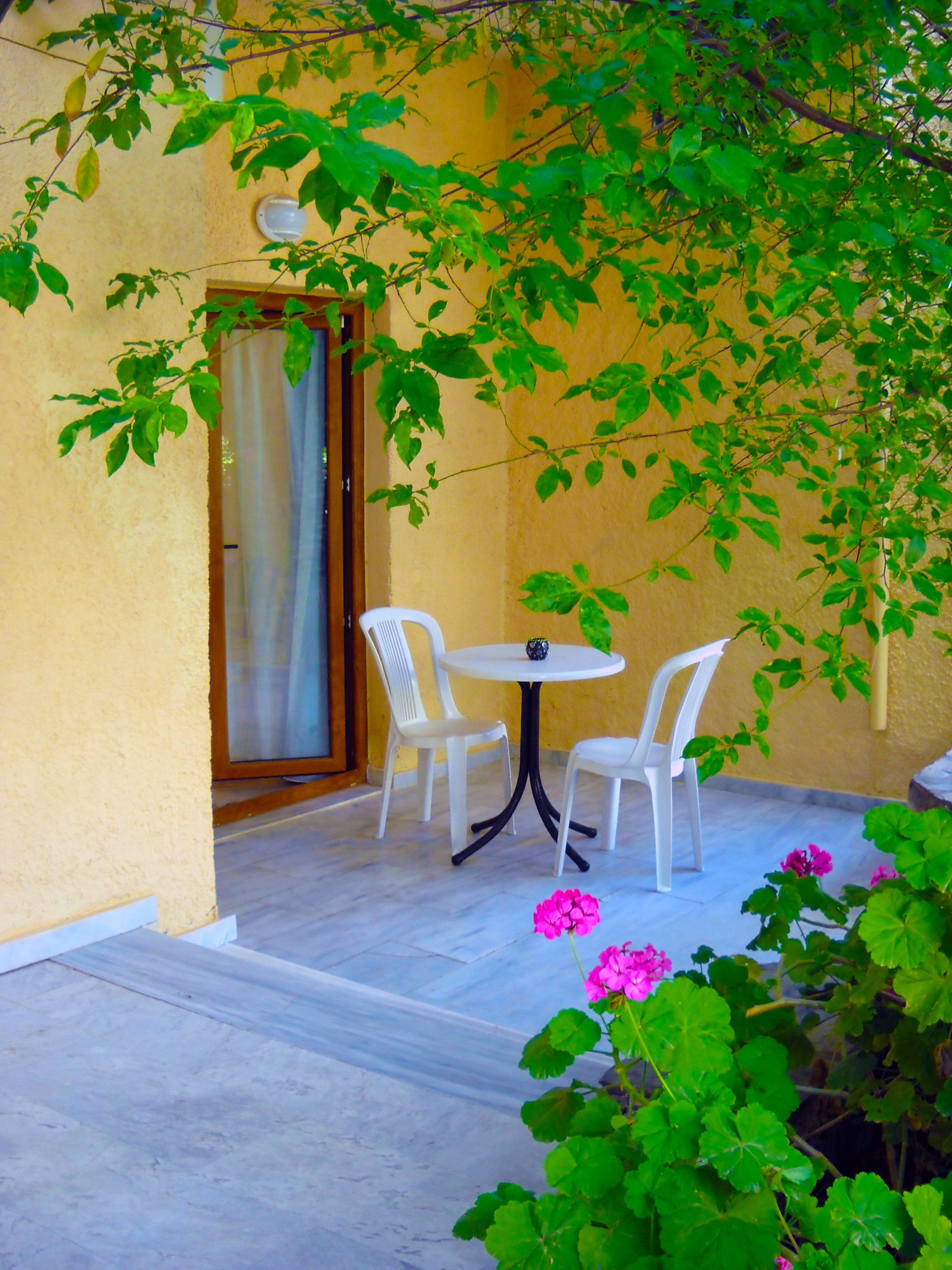A small outdoor seating area with two white chairs and a round table, surrounded by greenery and pink flowers.