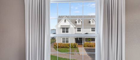 A window with white curtains, a view of a two-story house with a driveway and landscaping.