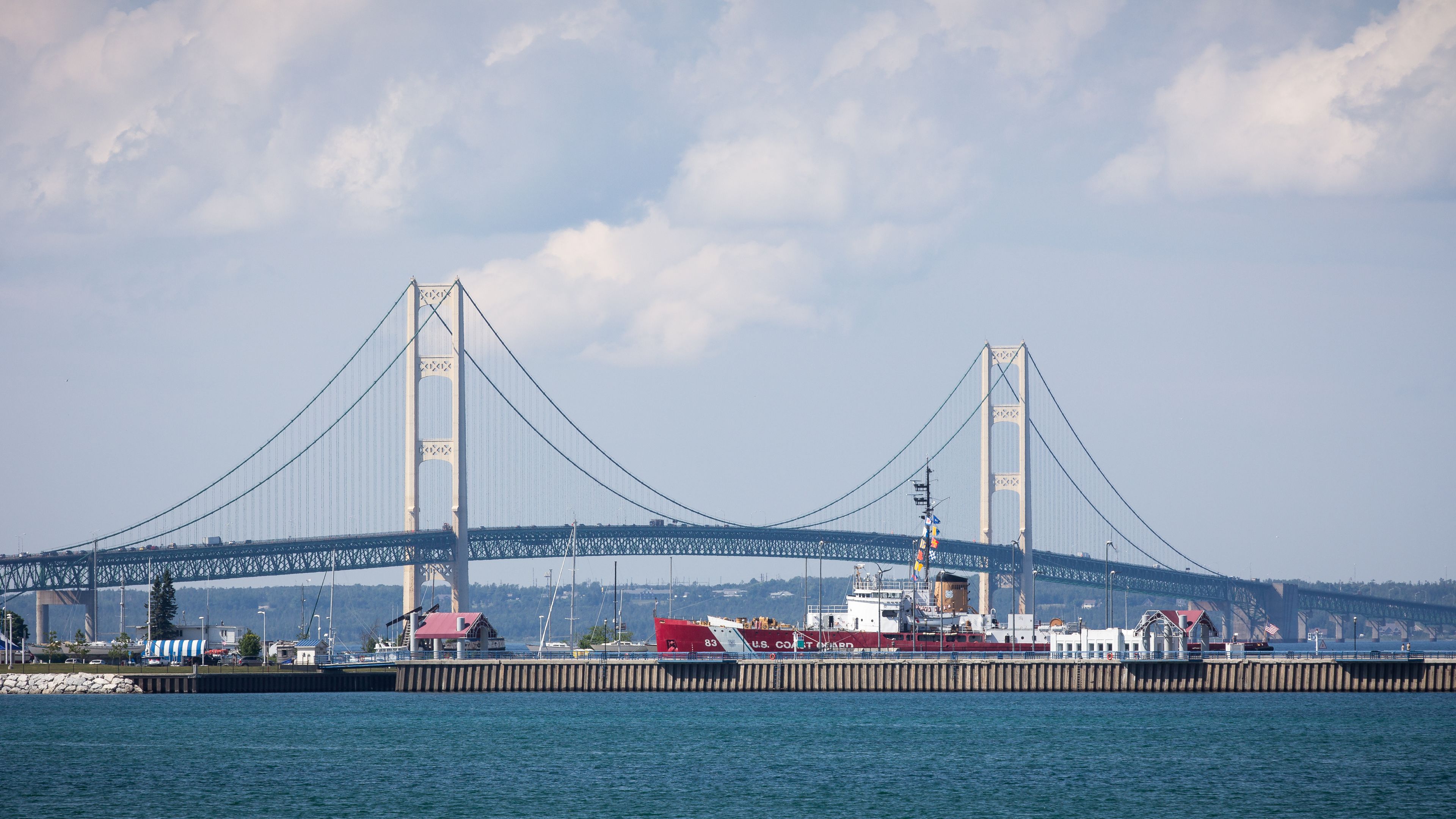A large suspension bridge with two towers and cables, a red ship docked at a pier, and a calm body of water in the foreground.