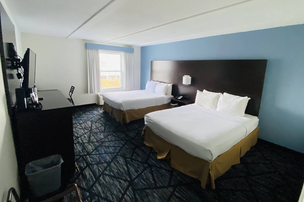 A hotel room with two beds, a desk, a chair, a trash bin, and a window with curtains.