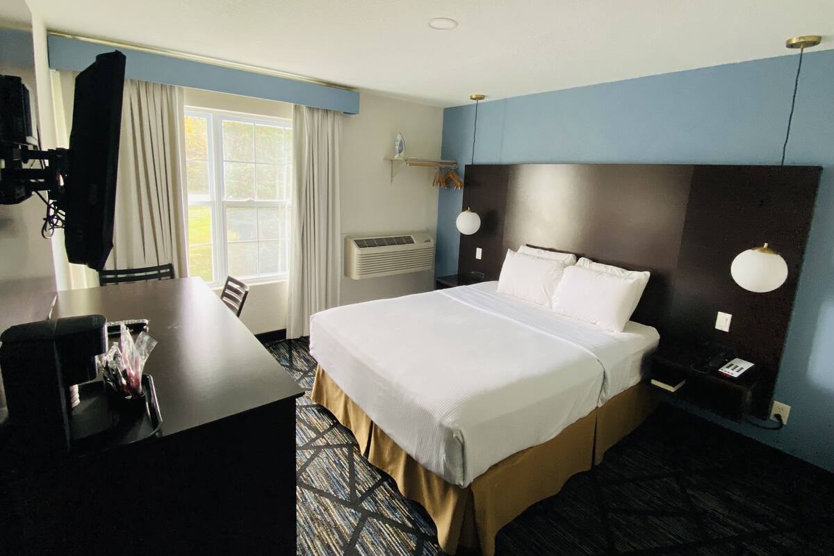 A hotel room with a bed, bedside tables, a desk, a chair, a window with curtains, an air conditioning unit, and a wall-mounted lamp.