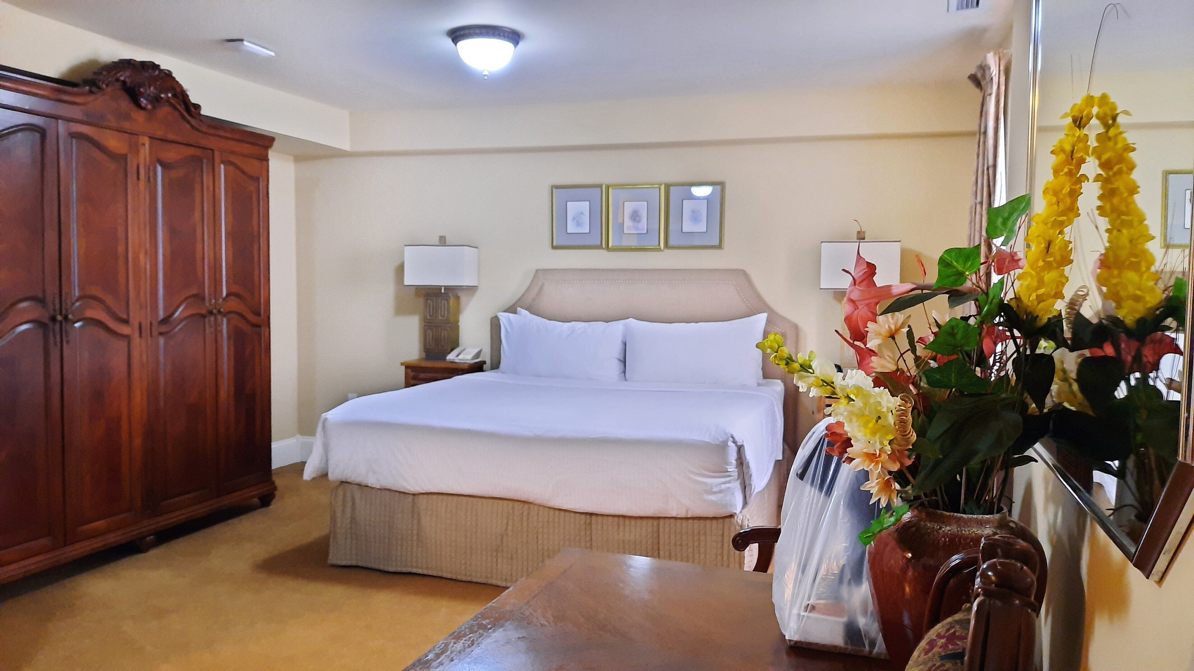 Habitación de hotel con una cama grande, un armario de madera, una mesita de noche con lámpara y un jarrón con flores.