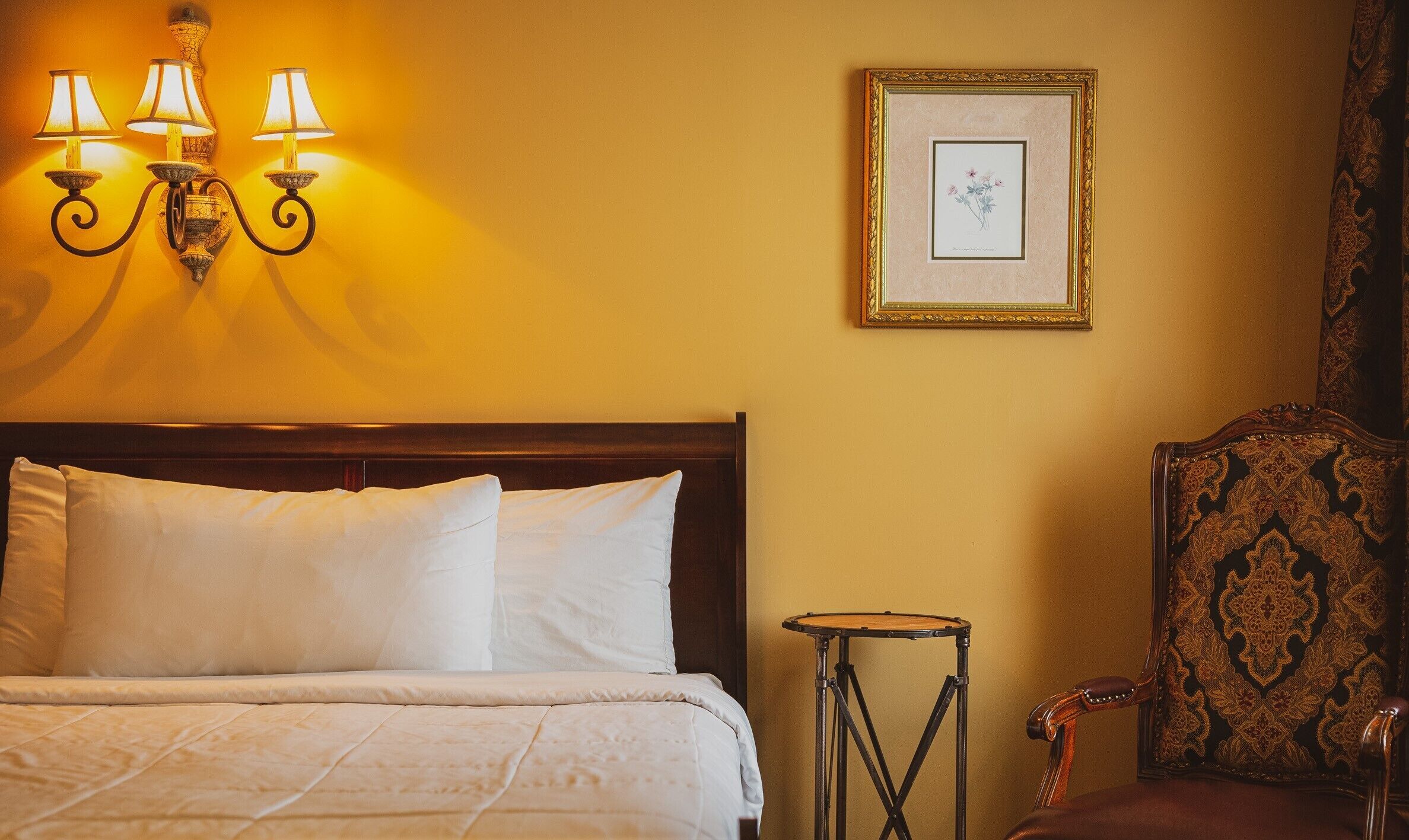 A neatly made bed with white linens, a wall-mounted lamp, a framed botanical print, and a patterned armchair.