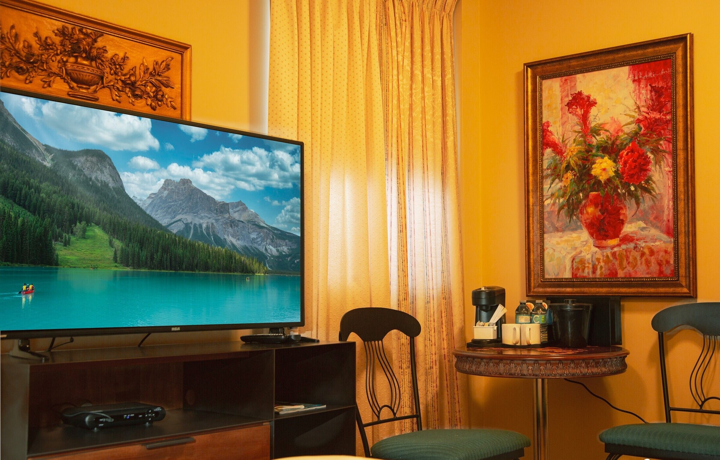 A living room with a large flat-screen TV displaying a mountain lake scene, a painting of flowers, and a small round table with a coffee maker.