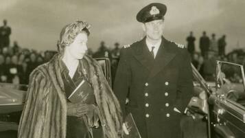 A historical scene with two individuals in military uniforms standing by a vehicle, with a crowd in the background.