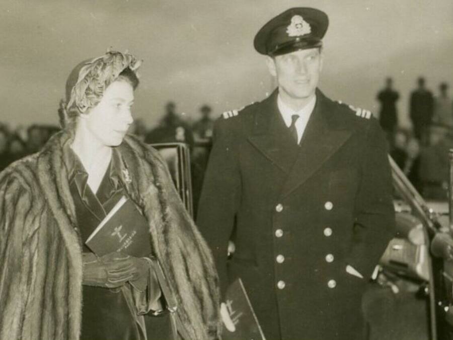 A historical scene with two individuals in military uniforms standing by a vehicle, with a crowd in the background.