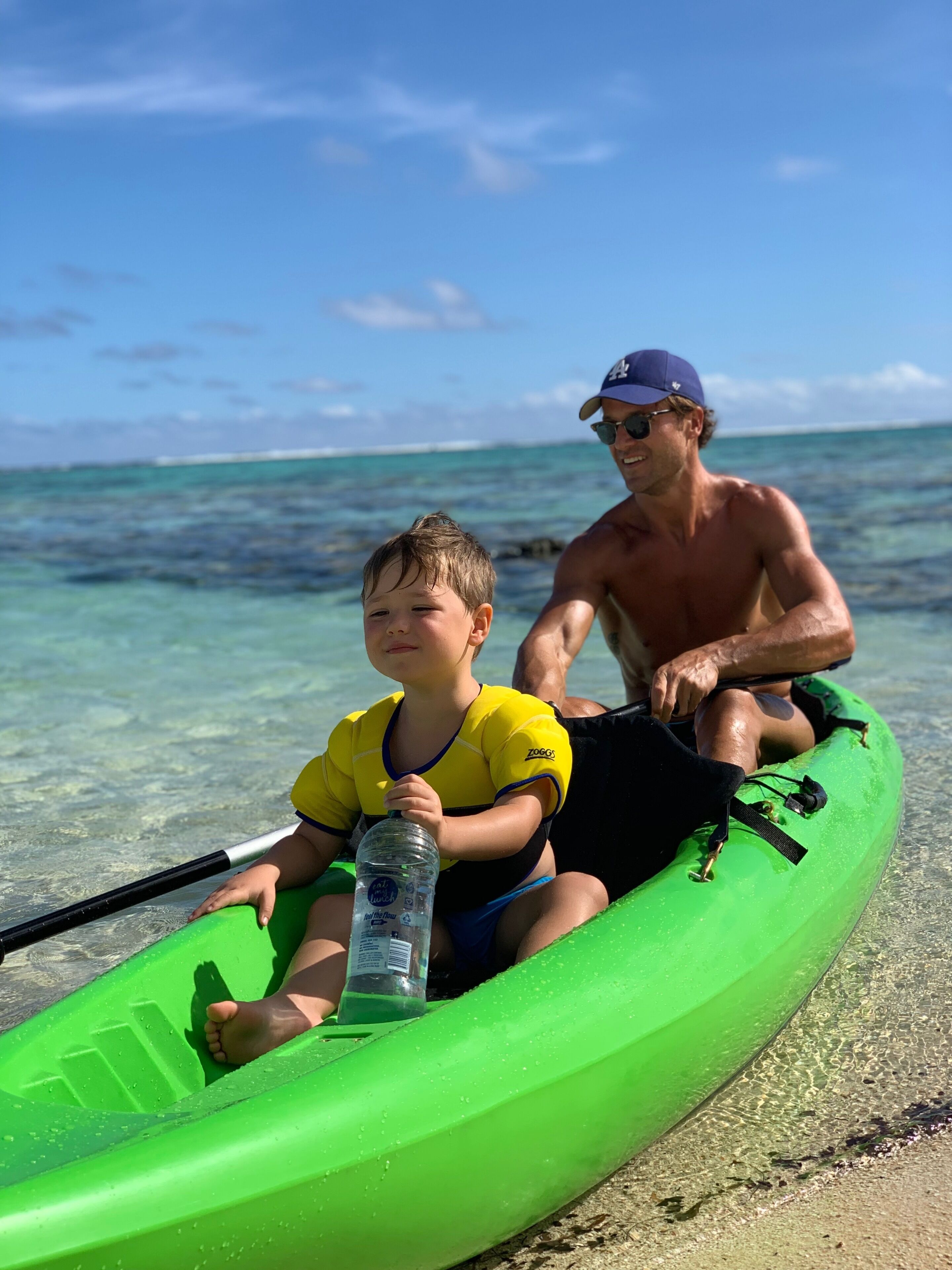 Un hombre y un niño en un kayak verde en una playa con agua cristalina y cielo azul.