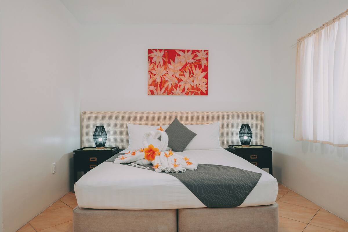 A neatly made bed with a white and gray bedspread, two nightstands with lamps, a red artwork on the wall, and a window with sheer curtains.
