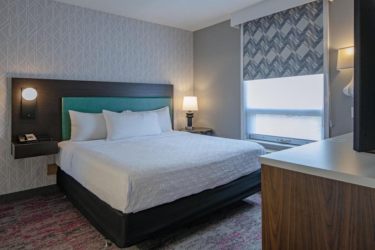 Habitación de hotel con cama, mesita de noche, lámpara y vistas al exterior.