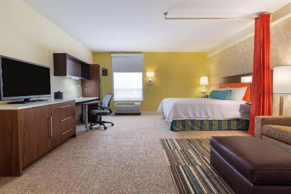A hotel room with a bed, a desk with a TV, a chair, a desk lamp, and a window with curtains.