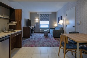 Habitación de hotel compacta con kitchenette, sofá, mesa de comedor y ventana con vistas.