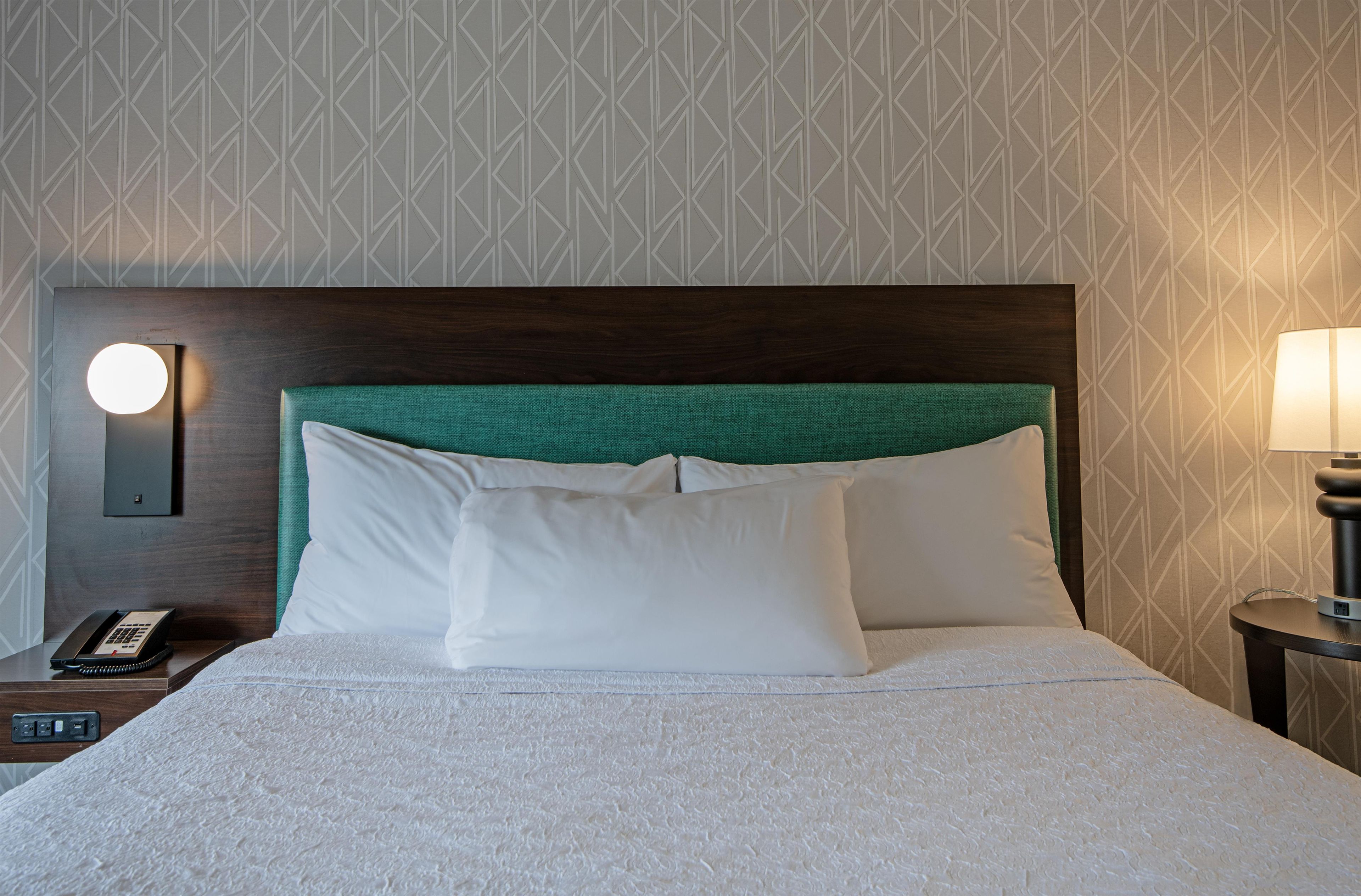 Ein Hotelzimmer mit einem ordentlich bezogenen Bett, einer Nachttischlampe, einem Telefon und einer Wandleuchte.