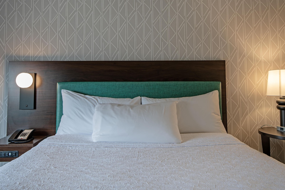 Habitación de hotel con una cama bien hecha, una lámpara de noche, un teléfono y un aplique de pared.