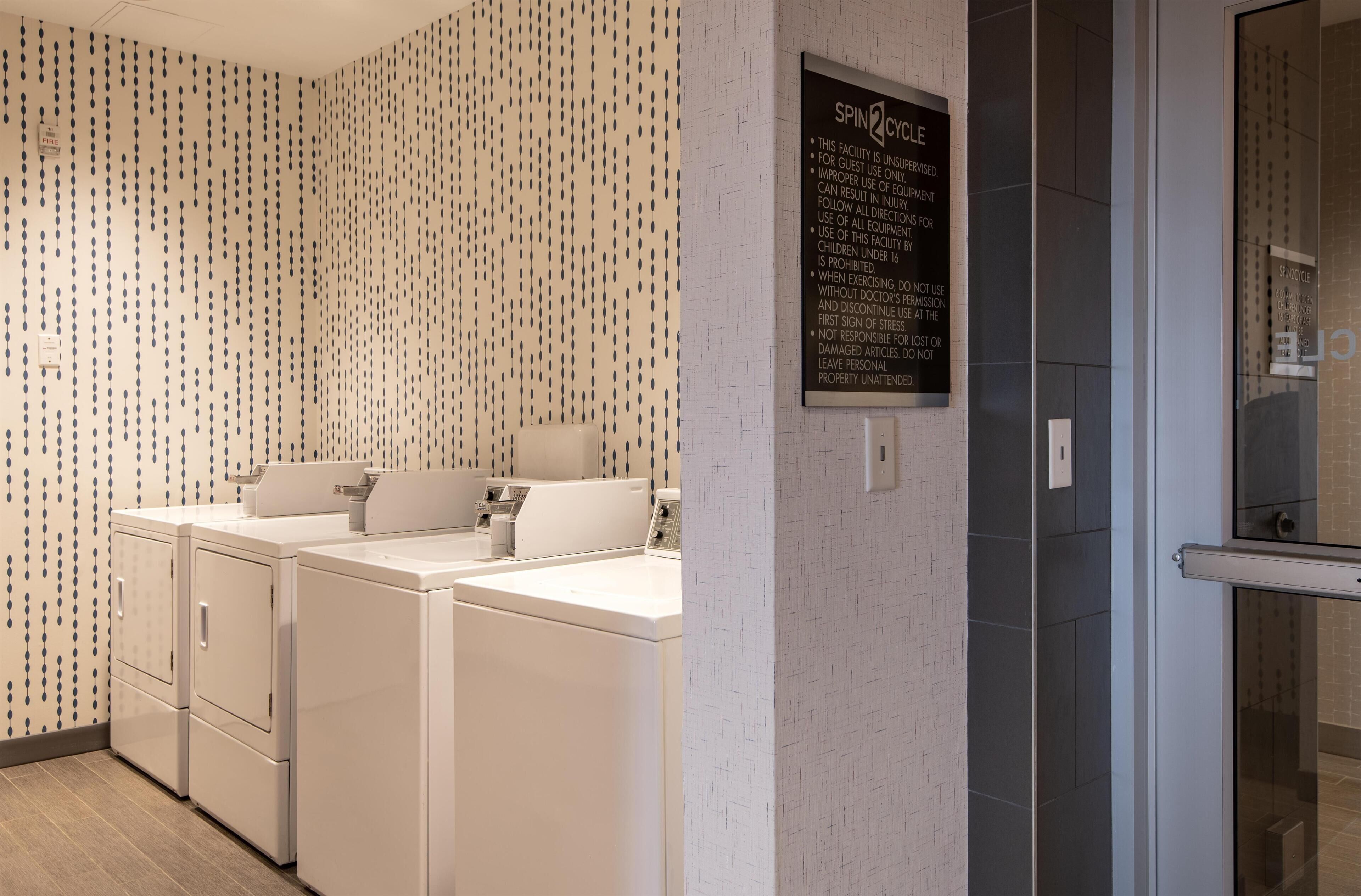 Une buanderie équipée de trois machines à laver et de trois sèche-linge blancs, un panneau affiché sur le mur et une porte en verre donnant accès à un espace douche.