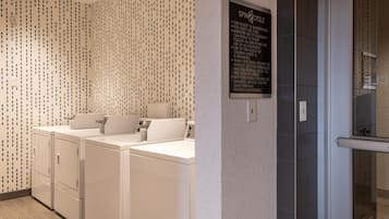 Une buanderie équipée de trois machines à laver et de trois sèche-linge blancs, un panneau affiché sur le mur et une porte en verre donnant accès à un espace douche.