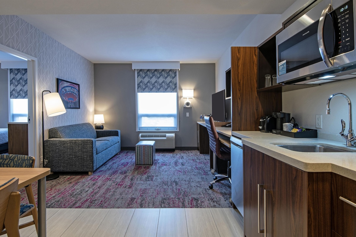Habitación de hotel compacta con kitchenette, sofá, mesa de comedor y ventana con vistas.
