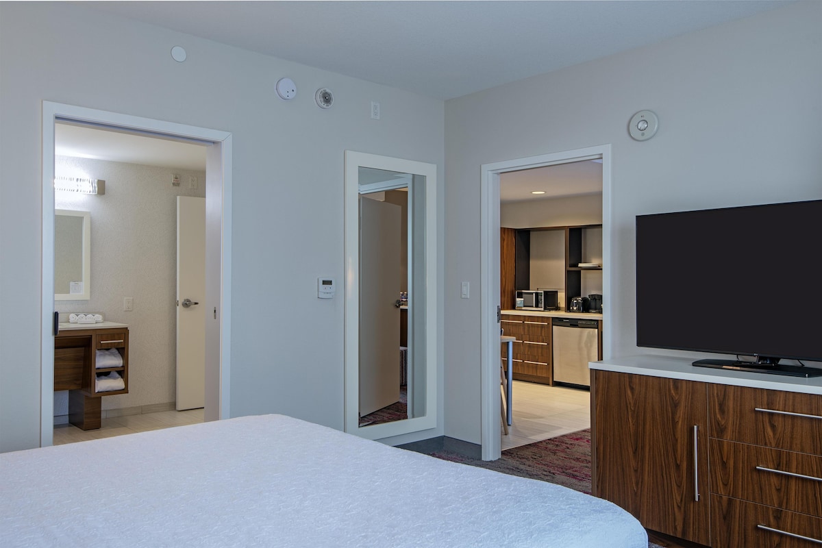 Habitación de hotel con cama, televisor, mueble de madera y cocina visible a través de una puerta abierta.