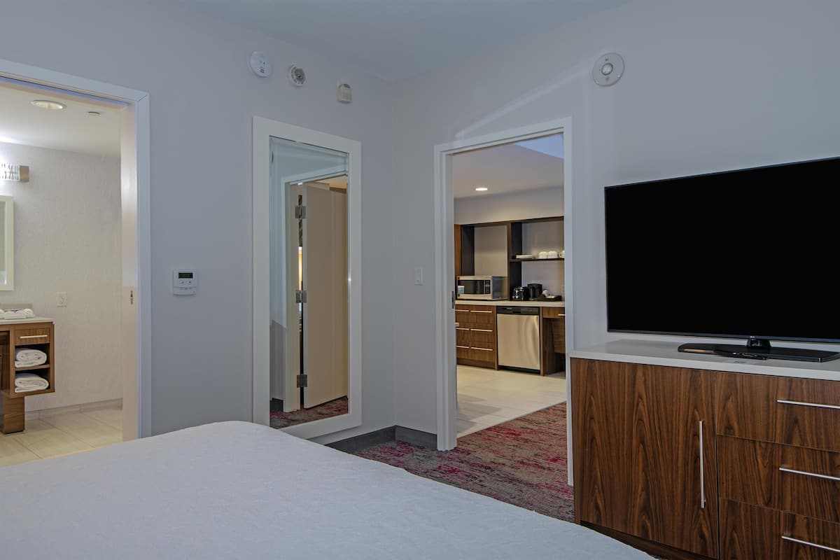 A hotel room with a bed, a TV, a wooden cabinet, and a kitchen area visible through an open door.