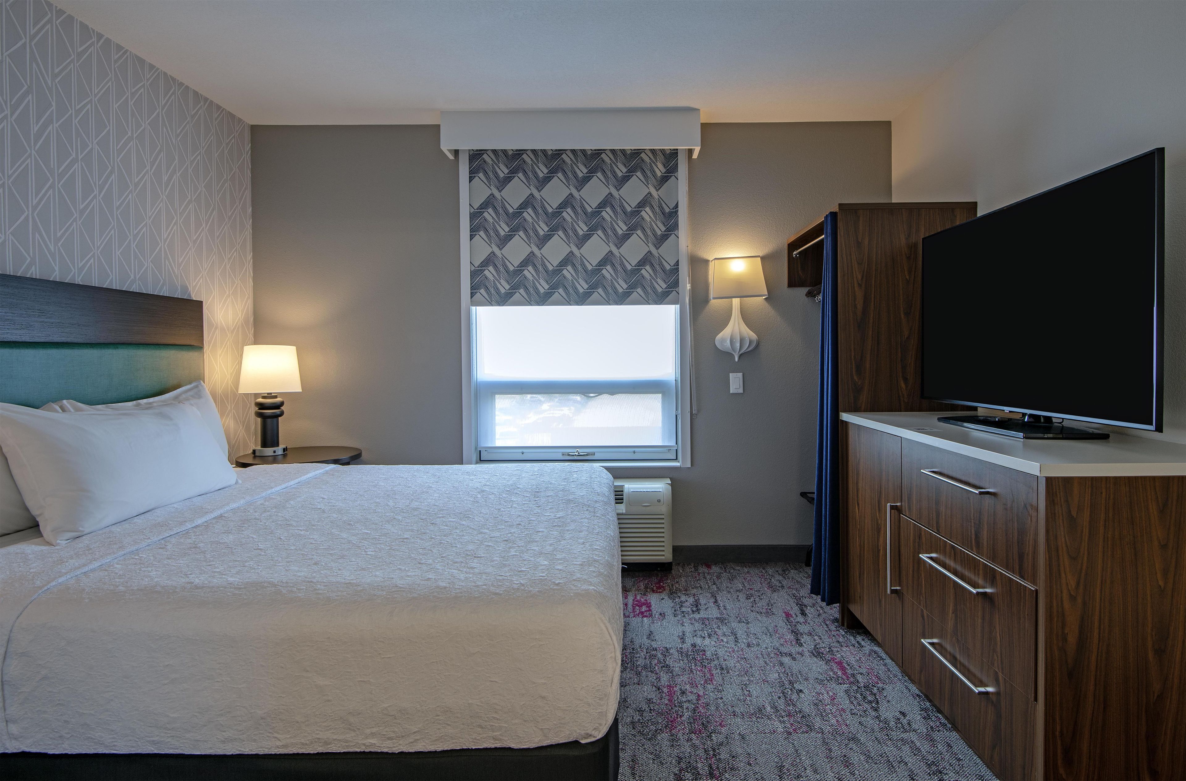 Habitación de hotel con cama, televisor, mesita de noche, lámpara, ventana con vistas y armario.
