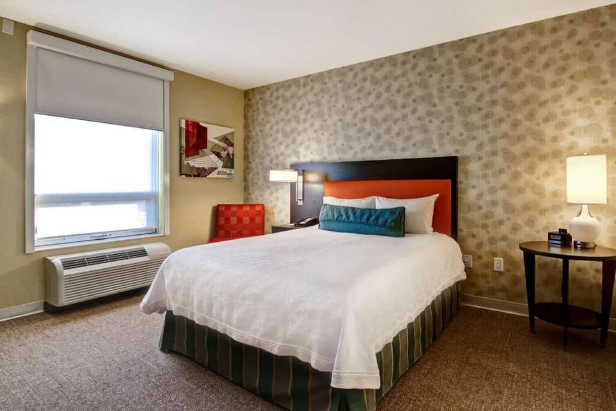 A hotel room with a bed, a window with blinds, a wall-mounted lamp, and a patterned wall.