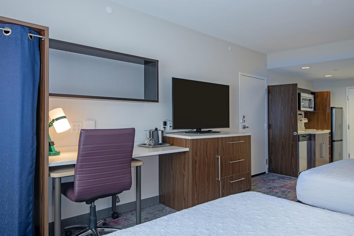 A hotel room with a bed, a desk with a computer, a TV, a kitchenette, and a shower area.