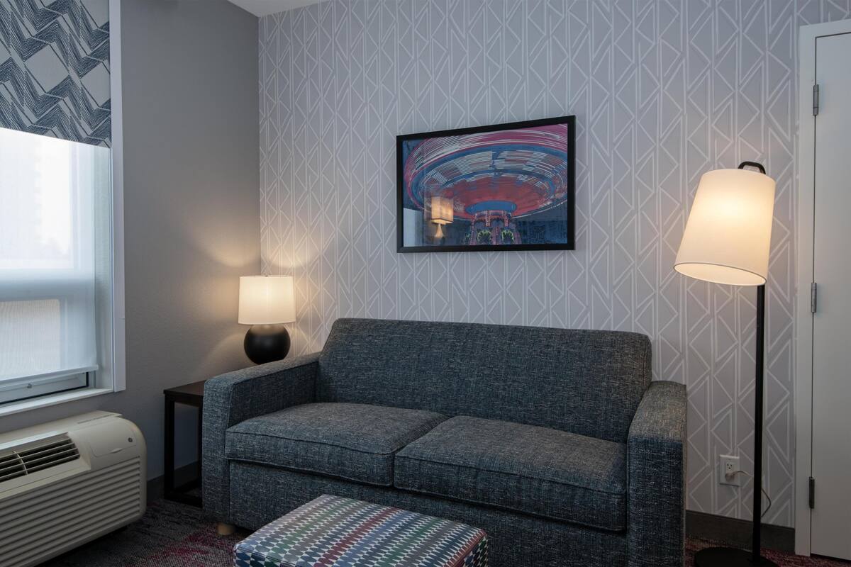 A room with a grey sofa, a patterned wall, a framed picture, a standing lamp, and a window with blinds.