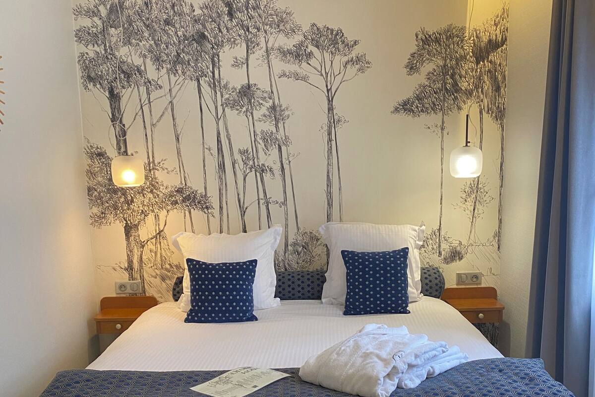 A neatly made bed with a patterned bedspread, two pillows with polka dots, and a towel on the bed. There is a wall with a nature-themed mural and two bedside lamps.