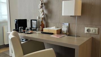 A hotel room with a desk, a chair, a flat-screen TV mounted on the wall, a lamp, a vase with flowers, a coffee maker, a bottle, a remote control, and a small box.