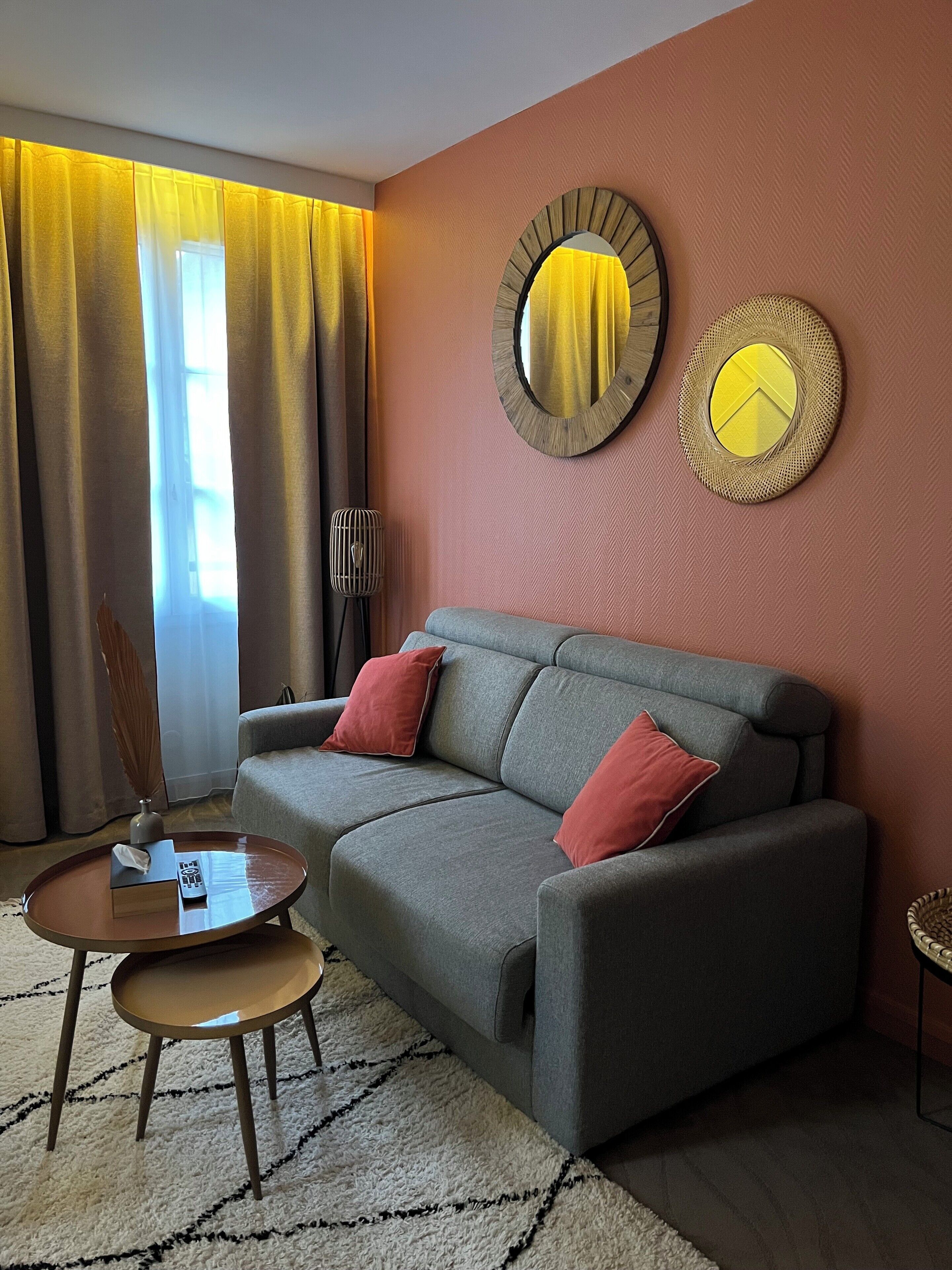 Una sala moderna con un sofá gris, dos cojines rojos, una mesa de centro redonda y una mesita auxiliar. Se observa un espejo grande con una cortina amarilla en la pared, y una lámpara de pie.