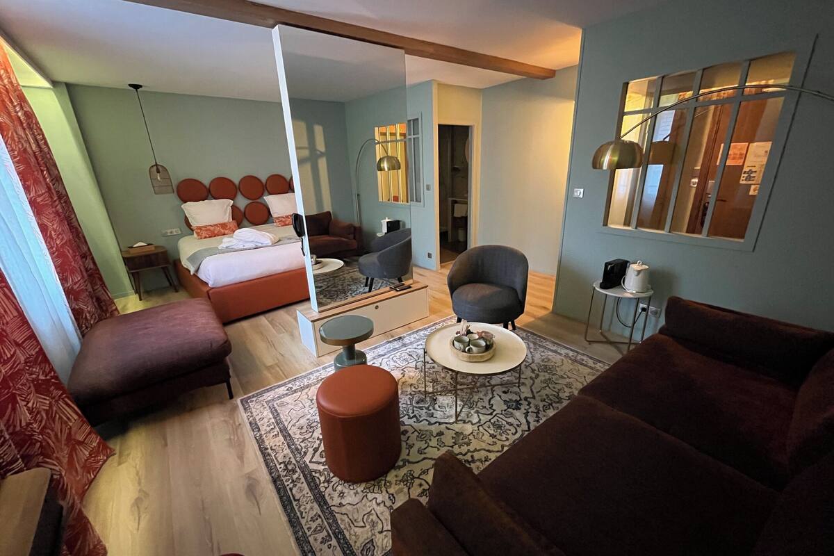 A modern hotel room with a bed, a sitting area with a sofa and armchair, a coffee table, and a large mirror.