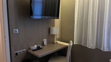 Habitación de hotel con televisor de pantalla plana montado en la pared, un escritorio con silla, una lámpara y una ventana con cortina blanca.