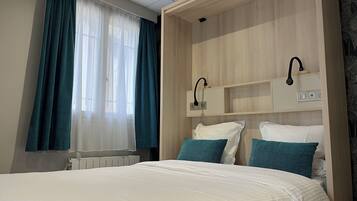 Une chambre d’hôtel avec un lit, une fenêtre avec des rideaux, une table de chevet et une étagère fixée au mur avec une lampe.