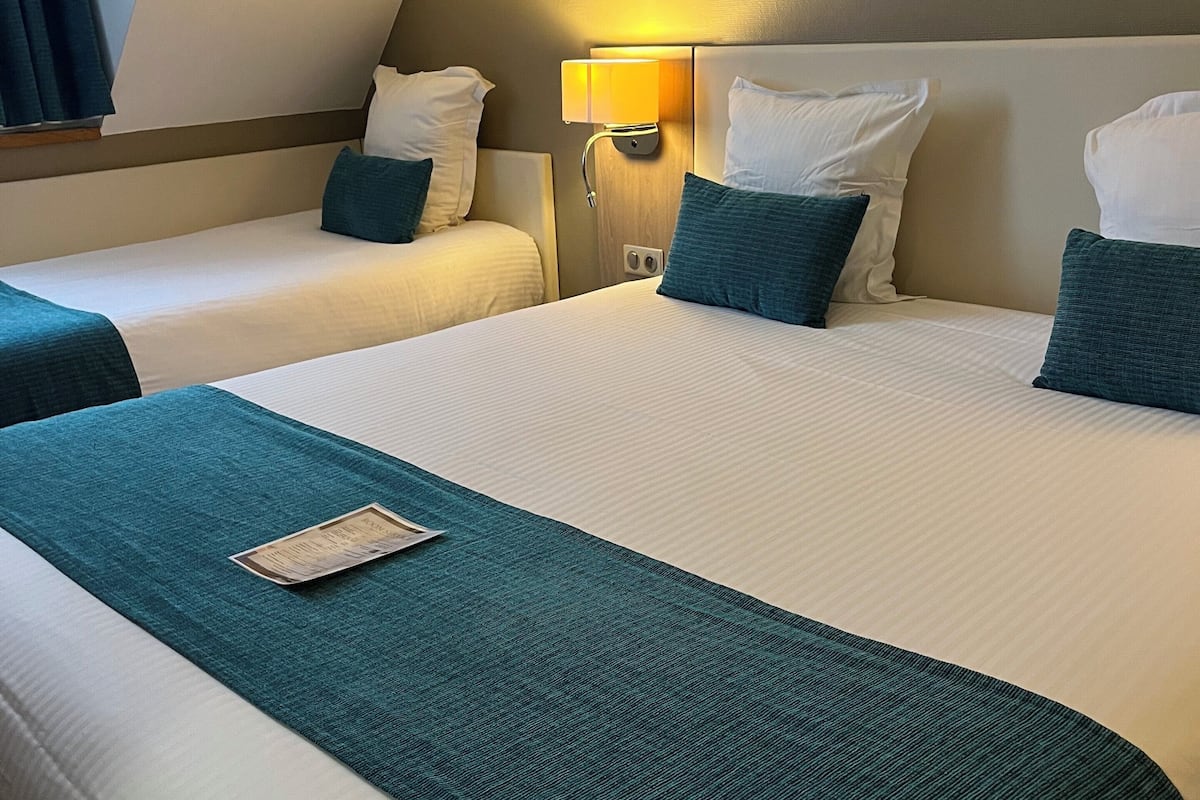 A hotel room with two beds, each with white linens and teal throw pillows. There is a bedside table with a lamp between the beds.